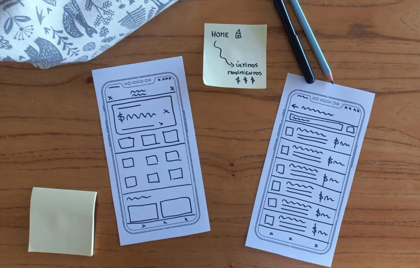 Dos bocetos a mano de pantallas de celular sobre una mesa de madera: el Home con la franja de saldo y los últimos movimientos, y el listado de movimientos con buscador, filtros y una fila por operación. Al lado, un post-it que dice «Home → Últimos movimientos» y dos lápices.